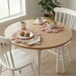 Two pink flower-shaped woven placemats on a round wood table with muffins, bread, coffee mugs, and white chairs.