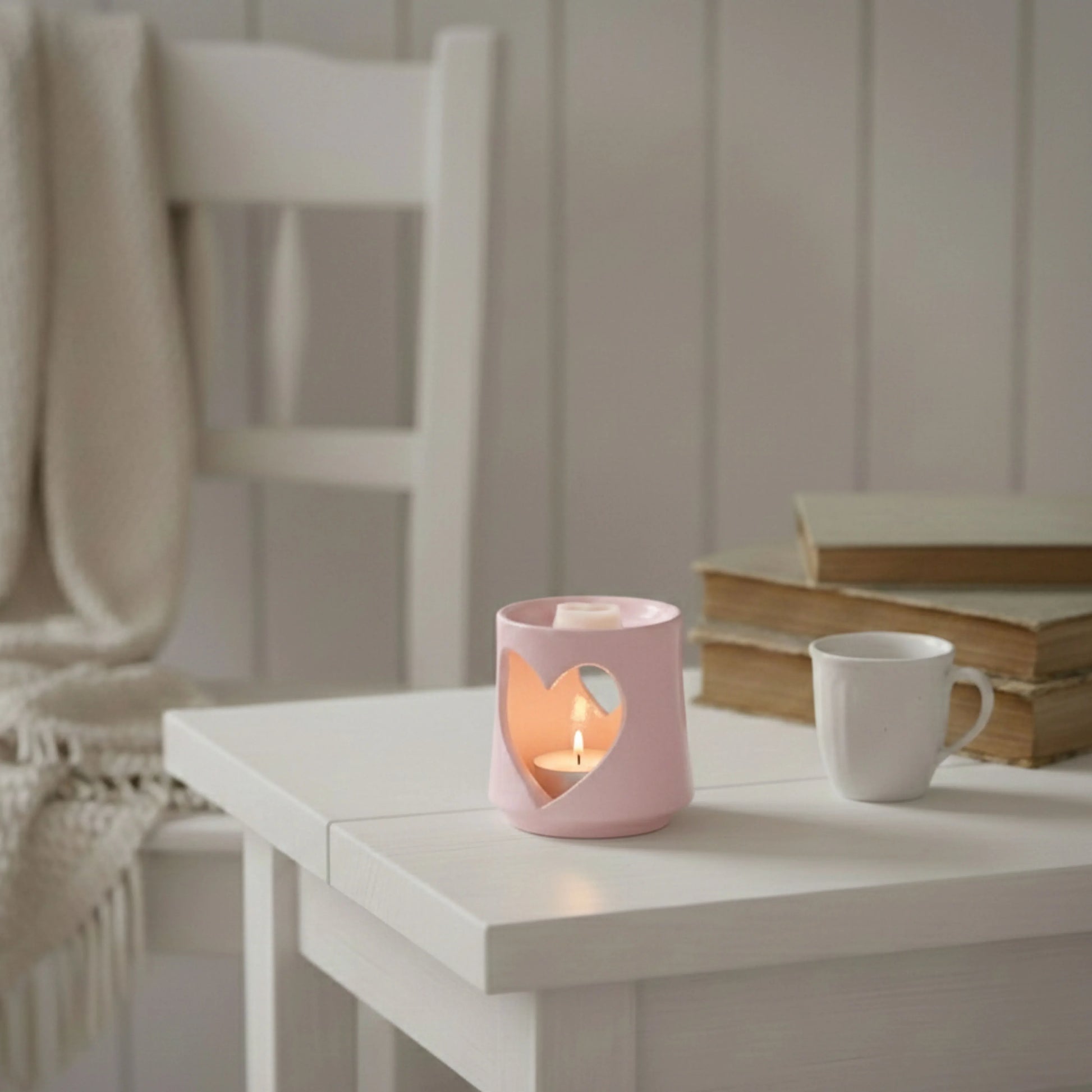 Pink heart-shaped candle holder with a lit candle on a white table, next to a white mug and books.

