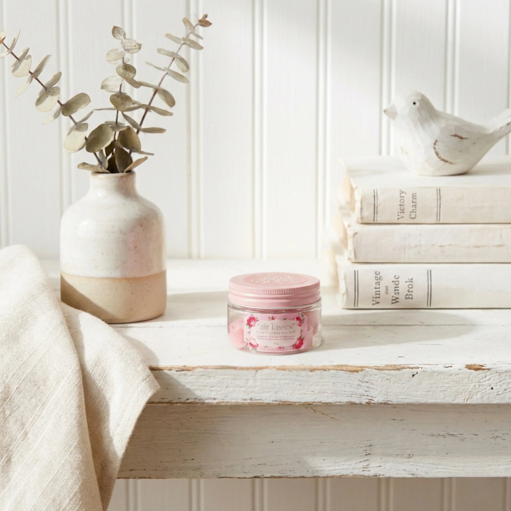 Pink jar on a wooden surface with a vase, books, and decorative items in the background.

