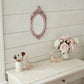 Pink ornate oval mirror on a white wood wall above a vanity with makeup brushes, perfume bottles, and pink peonies.