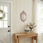 Pink ornate oval mirror on a shiplap wall over a wood table with a jute runner, pink roses, and a white Dutch door.