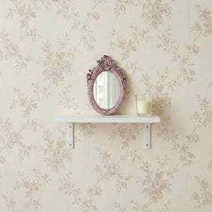 Small pink ornate oval mirror and a candle on a white shelf against floral rose wallpaper.