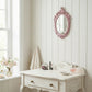 Pink ornate oval mirror on a white shiplap wall above a distressed wooden vanity with makeup and a perfume bottle.