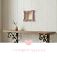 Small pink ornate mirror on a white wood wall above a rustic shelf with scrolled metal brackets and a pink heart icon.