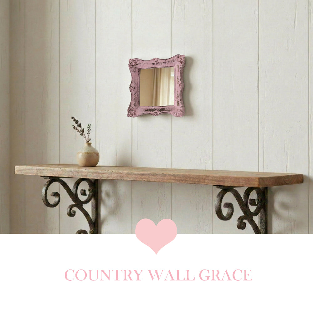 Small pink ornate mirror on a white wood wall above a rustic shelf with scrolled metal brackets and a pink heart icon.