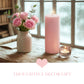 Lit pink pillar candle next to a vase of pink roses and a small tea light on a wooden surface by a window.