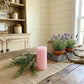 Pink pillar candle on a burlap runner with a metal tray of potted lavender and wooden beads on a rustic table.