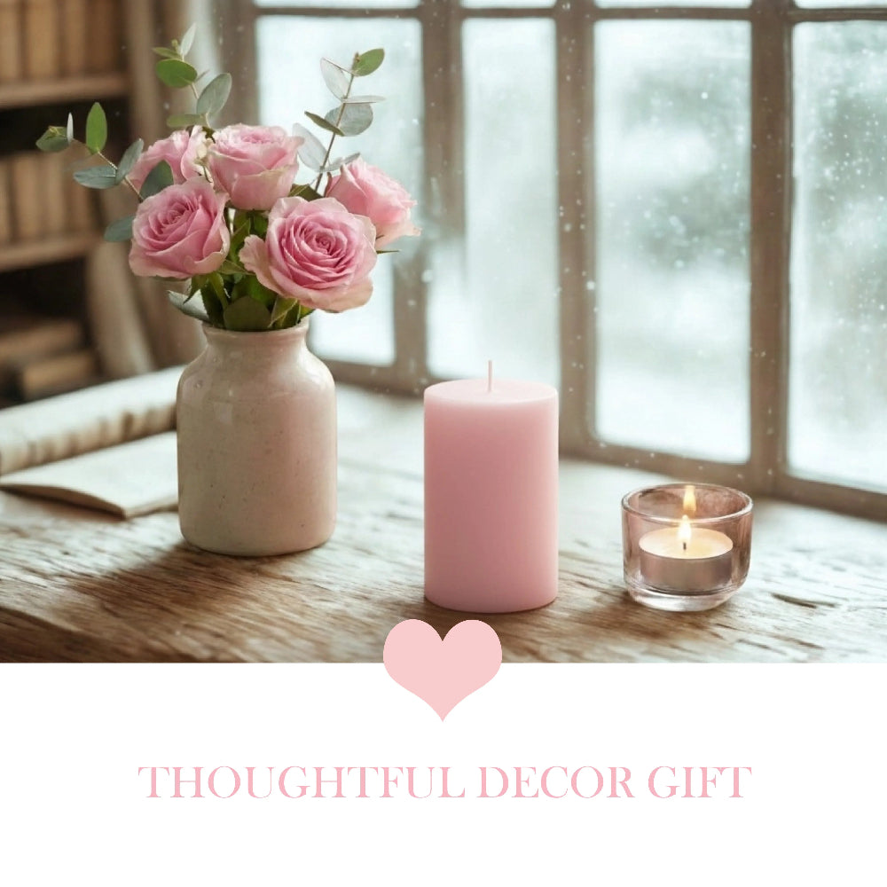 Pink pillar candle on a wooden windowsill next to a vase of pink roses and a small glass votive.