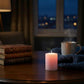 Lit pink pillar candle on a dark wood table with vintage books, a steaming mug, and a blue knit blanket near a window.