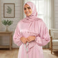 Woman wearing a pink hijab and polka dot Isdal Prayer dress in a bright room with a plant and chair.