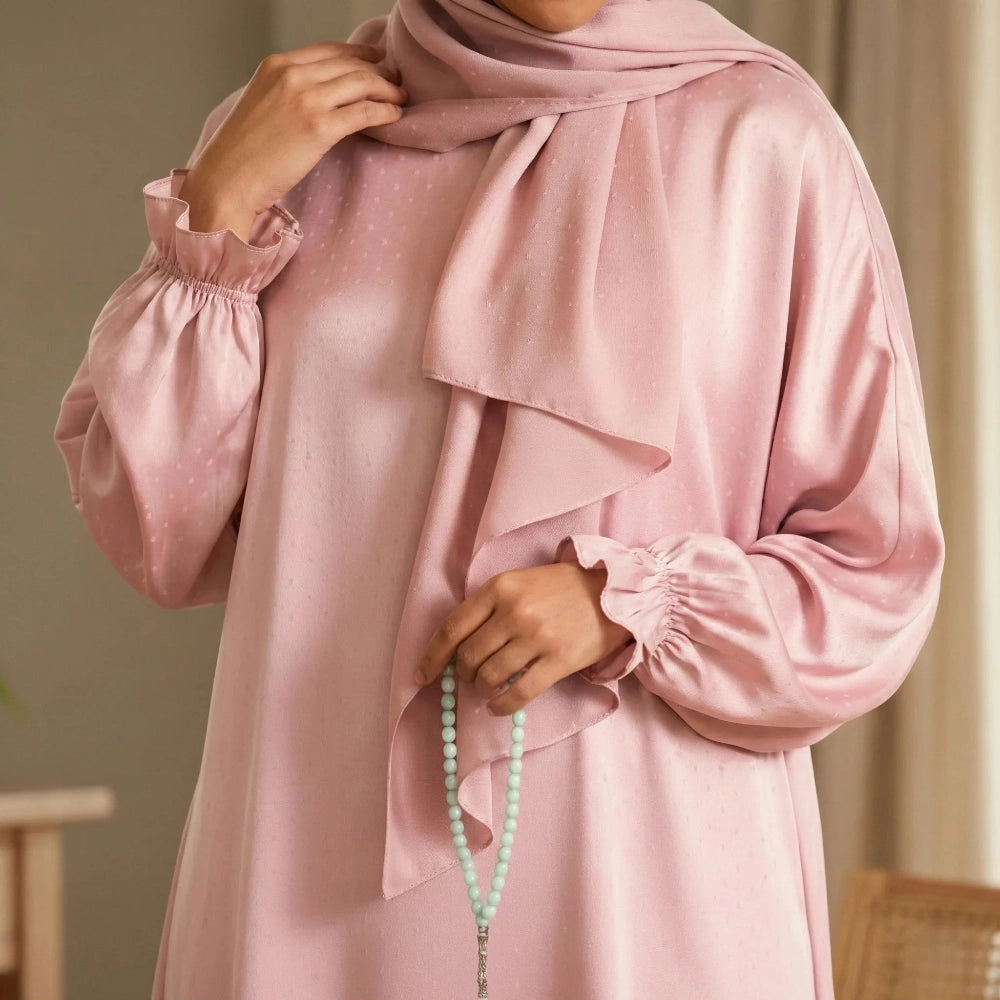 Person wearing a polka dot Isdal Prayer dress with a hijab, holding a rosary, in a neutral indoor setting.
