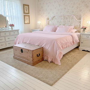 Pink ruffled bedding on a white metal bed frame with a wooden chest, tan rug, and white furniture in a bright bedroom.