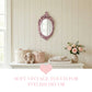 Pink ornate mirror above a white dresser with fabric bins, a plush rabbit, folded linens, and roses in a vase.
