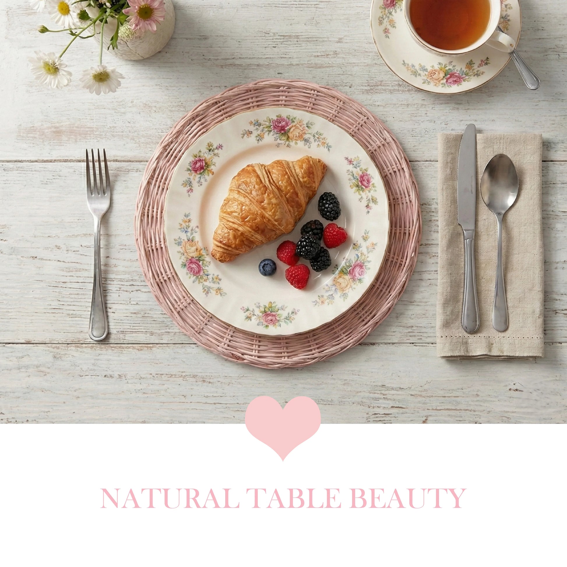 Breakfast setting with a croissant and berries on a floral plate, pink wicker placemat, and tea on a white wooden table.