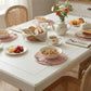 Breakfast table with pink wicker placemats, bowls of granola, sliced bread, a teapot, and flowers on a white table.