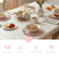 Breakfast table with pink wicker placemats, bowls of granola, fruit, a teapot, and bread on a white wooden table.