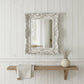 Decorative mirror with ornate frame above a wooden shelf on a white paneled wall.