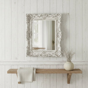 Decorative mirror with ornate frame above a wooden shelf on a white paneled wall.