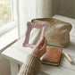 Pretty Shabby Wall Mirror – Rectangular Chic held in hand above beige pouch on wooden surface near window