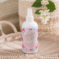 Country Charm fragrance mist bottle on a woven surface with flowers in the background