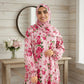 Woman wearing a pink floral dress and hijab in a room with wooden walls and a table with flowers.