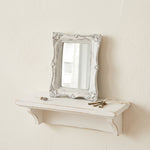 Ornate rectangular antique white mirror on a distressed floating shelf with a small flower and vintage keys.