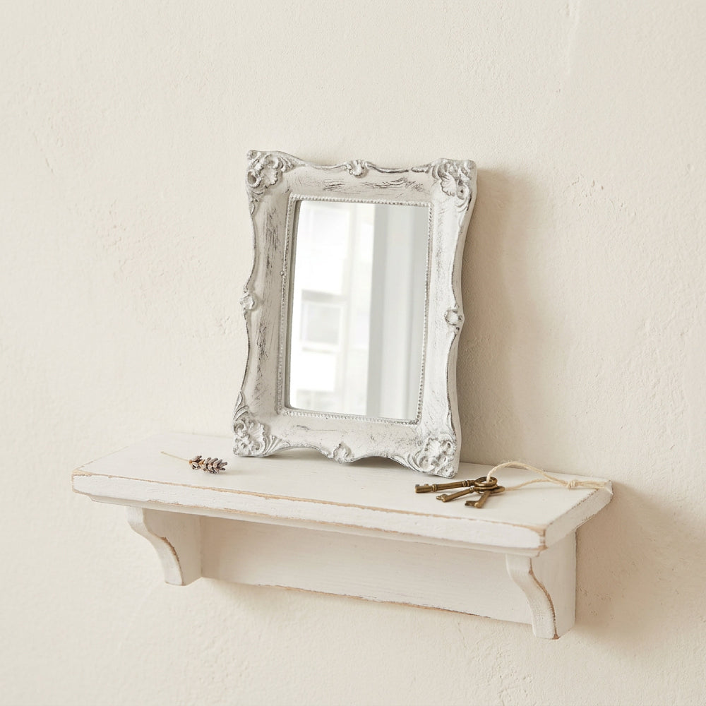 Ornate rectangular antique white mirror on a distressed floating shelf with a small flower and vintage keys.
