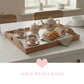 Wooden floral tray with a tea set, croissants, and cream pitcher on a white table, featuring a pink heart below.