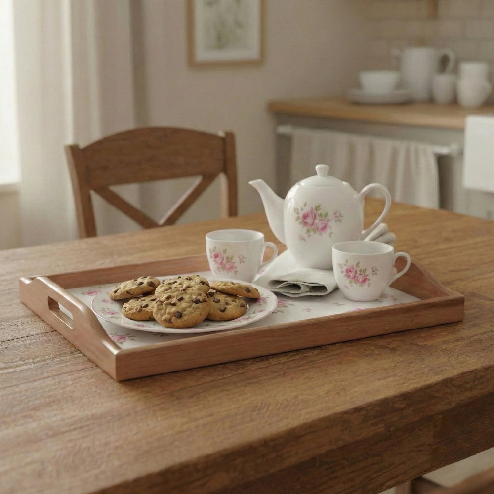 Wooden tray with floral patterns holding a teapot, two teacups, and a plate of chocolate chip cookies on a wood table.