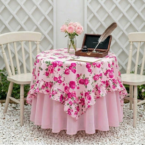 Round table with pink floral tablecloth, vase of flowers, and writing set outdoors.