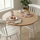 Round wooden table with white chairs, coffee, pastries, and a loaf of bread in a bright room.
