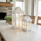 Two white decorative lanterns with warm glowing fairy lights inside, placed on a light wood dining table in a bright living room setting.