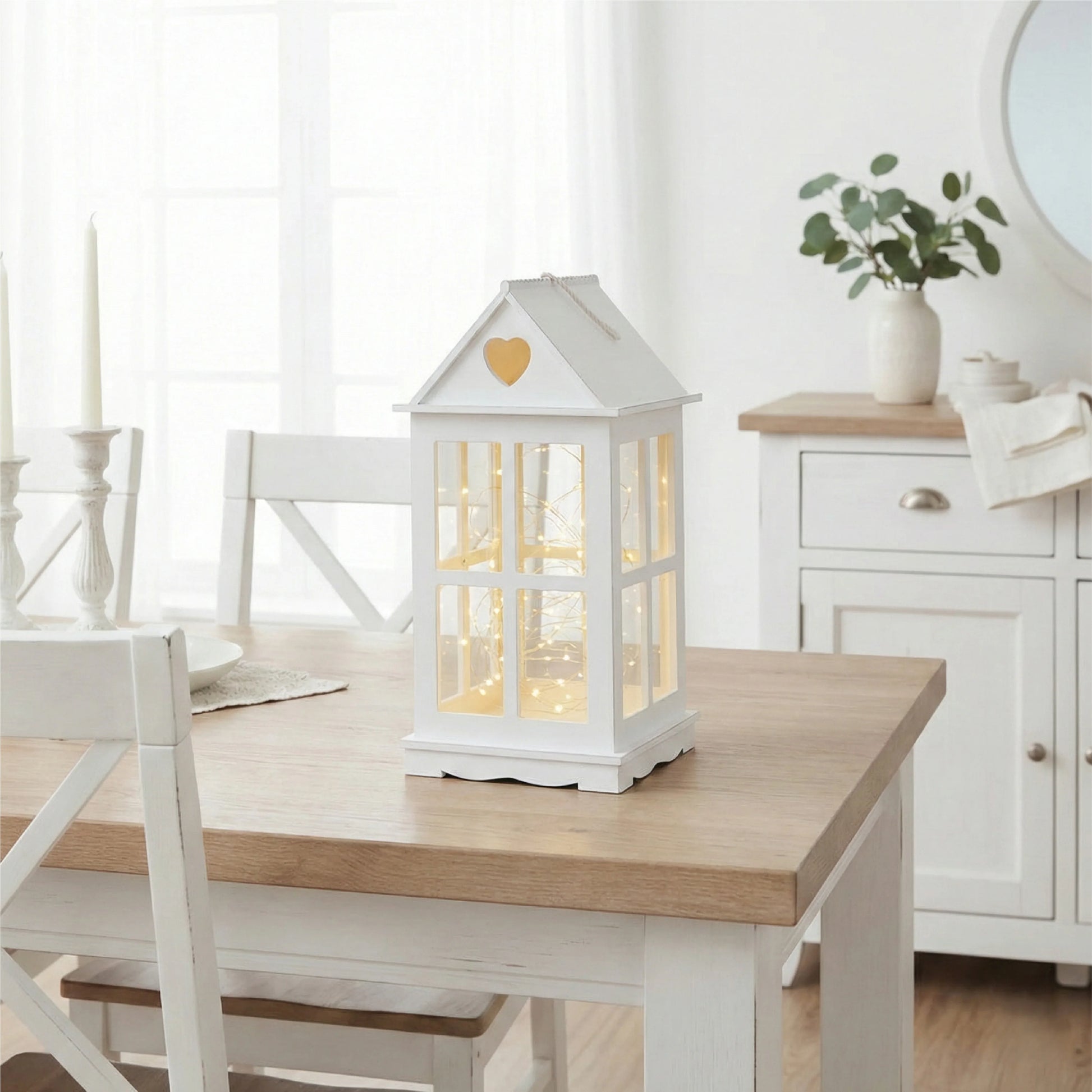 White house-shaped lantern on a wooden table in a bright room with white walls and furniture.