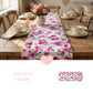 Pink rose floral table runner with ruffled edges on a wooden dining table set with white teacups and a vase of roses.