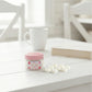 Small jar with pink lid on a white surface with a cup and book in the background

