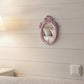 Small ornate pink mirror on a white shiplap wall reflecting a bed with pillows.