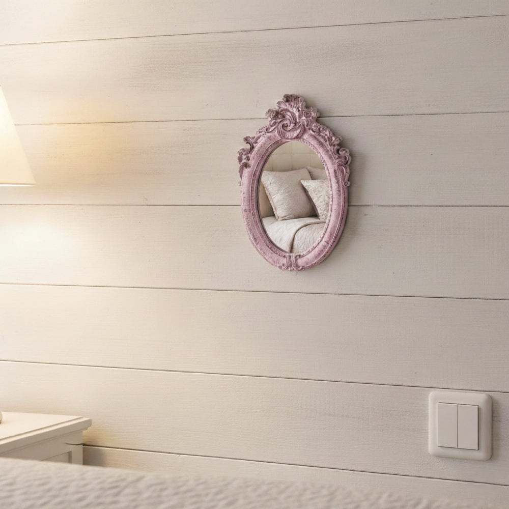 Small ornate pink mirror on a white shiplap wall reflecting a bed with pillows.