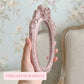 Hand holding a small pink ornate oval mirror in front of floral wallpaper and a vintage chair.