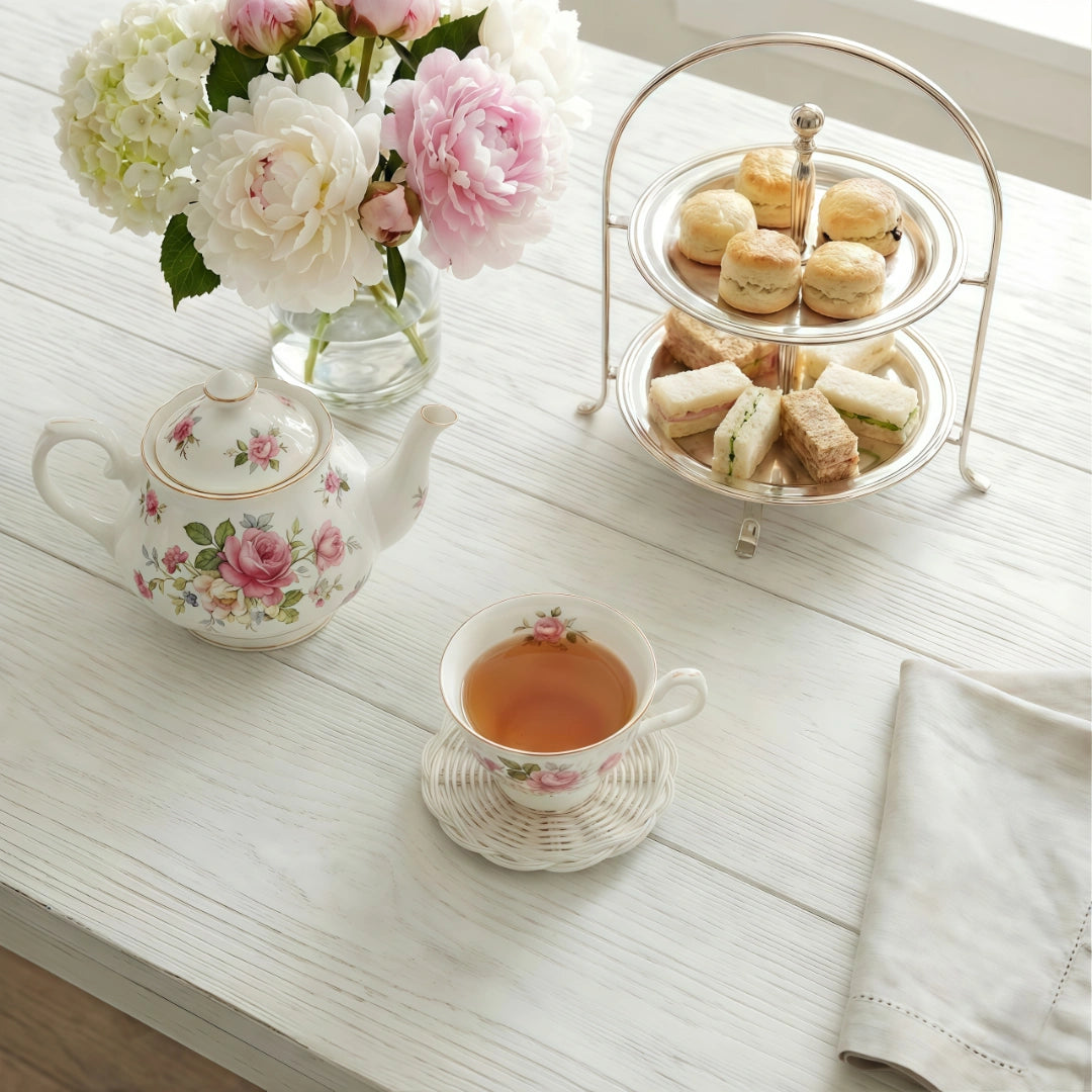Tea set with teapot, cup, and tray of sandwiches on a wooden table with flowers.
