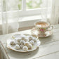 Teacup with kahk treat on a white wooden table near a window with lace curtains.
