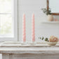 Two pink twisted candles in glass holders on a wooden table with a blurred background