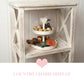 Two-tier white heart tray holding chocolates and candies on a distressed wooden shelf with pink text.