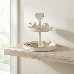 Two-tier antique white tray with a heart handle on a wooden shelf, holding small bird figurines and wooden beads.