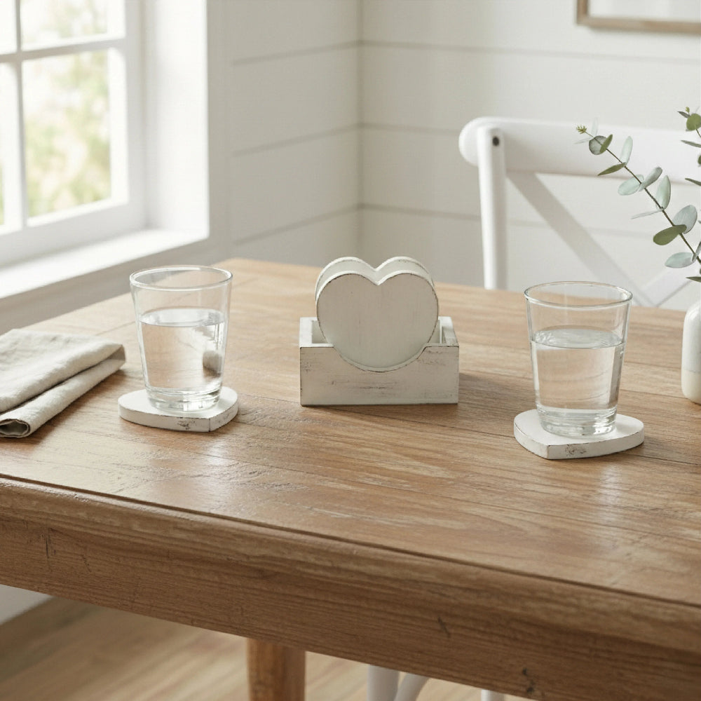Two glasses of water on heart coasters next to a wooden coaster holder on a natural wood table by a window.