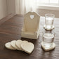 Two glasses of water on heart coasters next to an empty tall white wooden holder on a dark wood table.