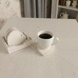 White coffee cup on a heart-shaped coaster next to a matching coaster set on a light wood table with floral wallpaper.