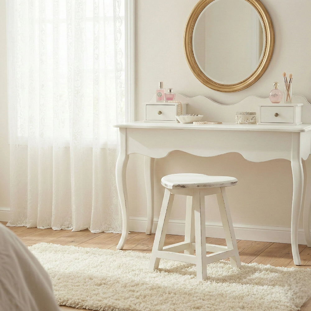 White heart-shaped stool in front of a curved vanity desk with a gold round mirror and perfume bottles.