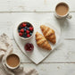 Top-down view of a white heart-shaped wooden tray with two croissants, a bowl of berries, and jam, served with two cups of coffee on a distressed white wood background.