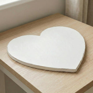 White heart-shaped wooden tray with a distressed finish resting on a light wood surface near a window.