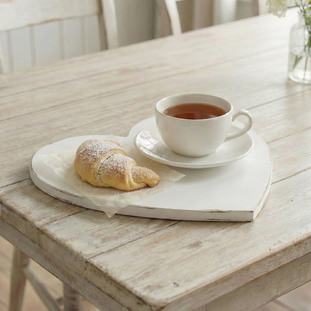 White heart-shaped wooden tray with a distressed antique finish, holding a powdered croissant on parchment paper and a white ceramic cup of tea on a rustic whitewashed table.